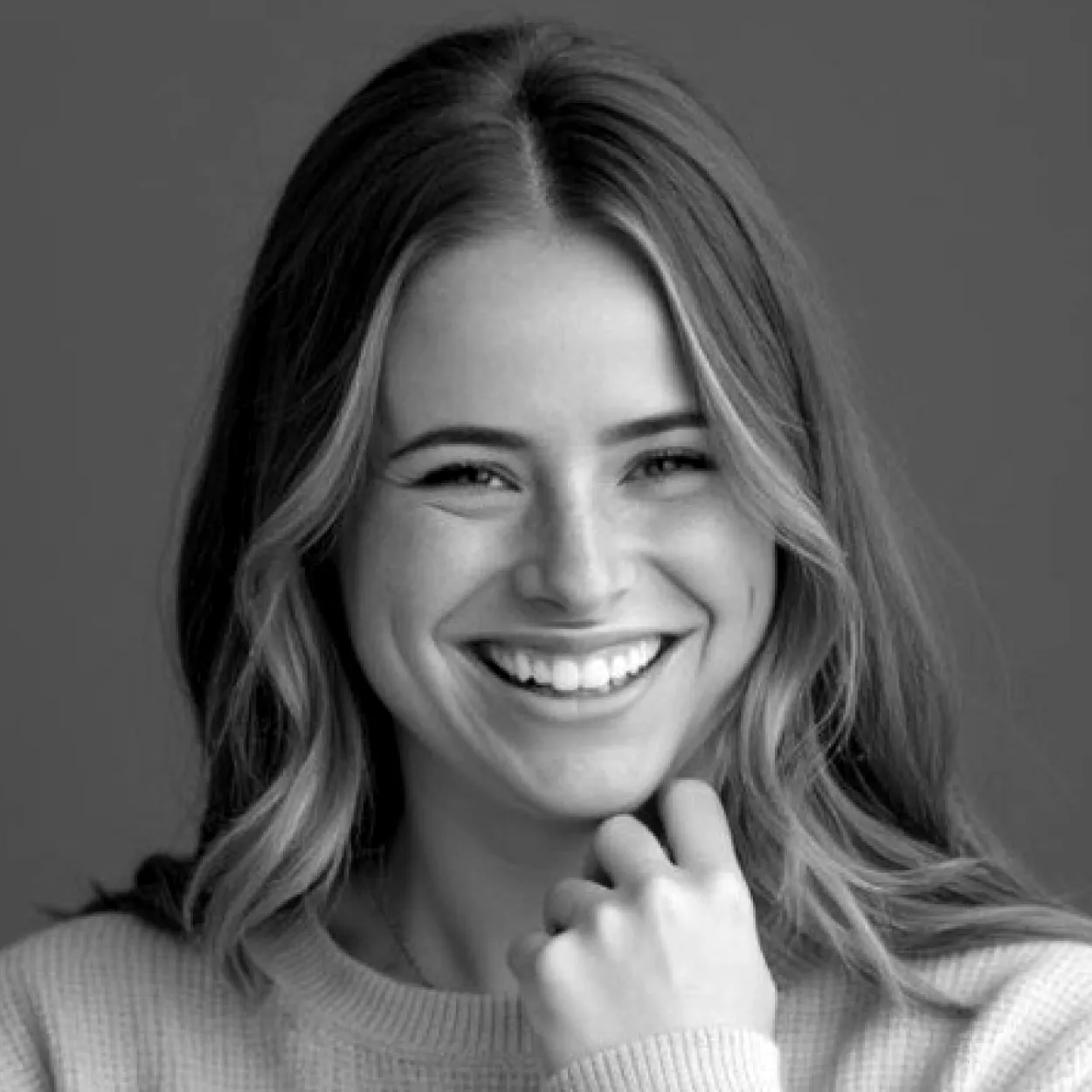 Smiling woman with long hair and a light sweater resting her fist near her chin.