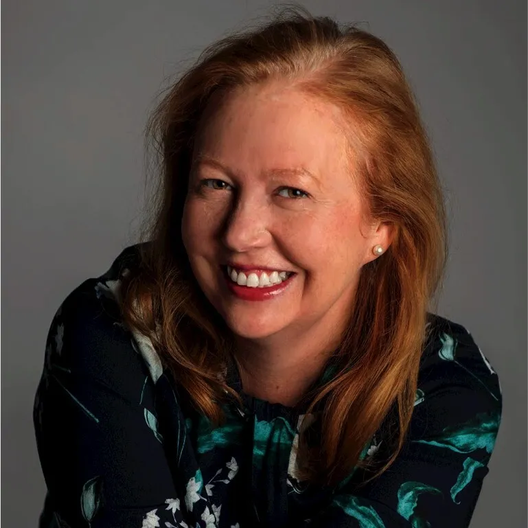 Smiling woman with red hair wearing a dark floral blouse against a gray background.