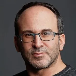 Man with short dark hair, glasses, and light facial hair wearing a dark shirt against a gray background.