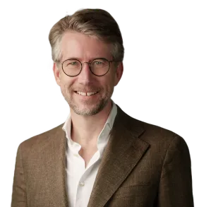 Smiling man with light brown hair and glasses wearing a brown blazer over a white shirt.