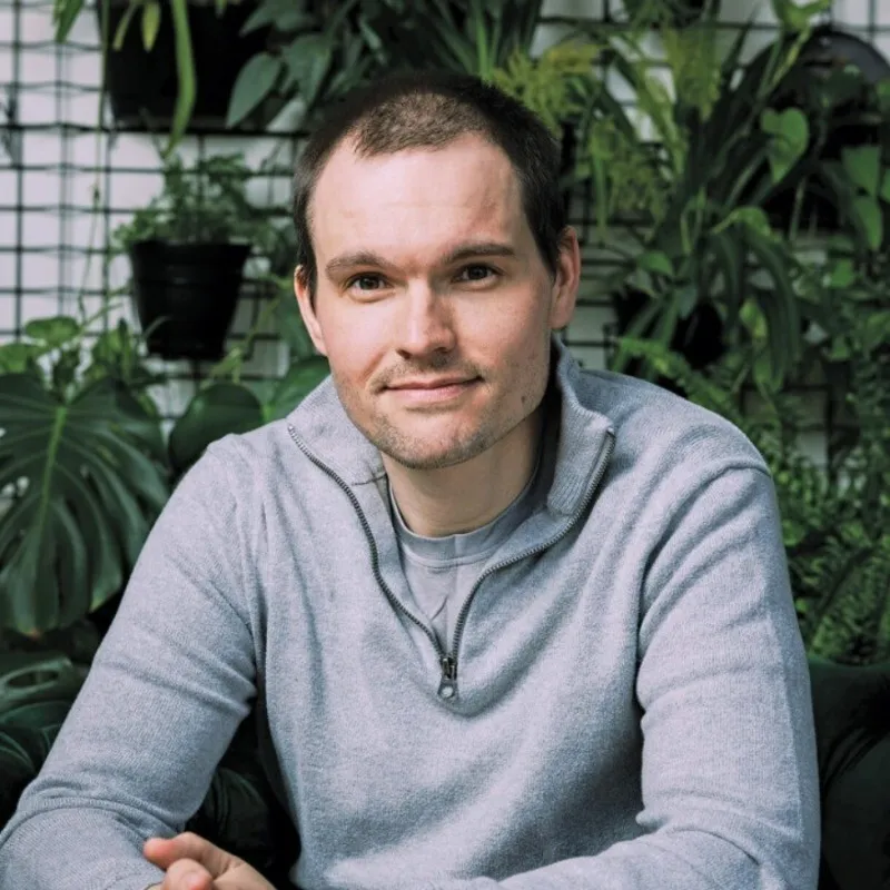 Man with short brown hair wearing a light gray quarter-zip sweater sitting in front of a green leafy background.