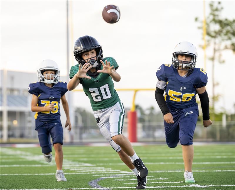 Youth football players on a field during a game, with one player in a green jersey catching a football while two players in blue jerseys chase.