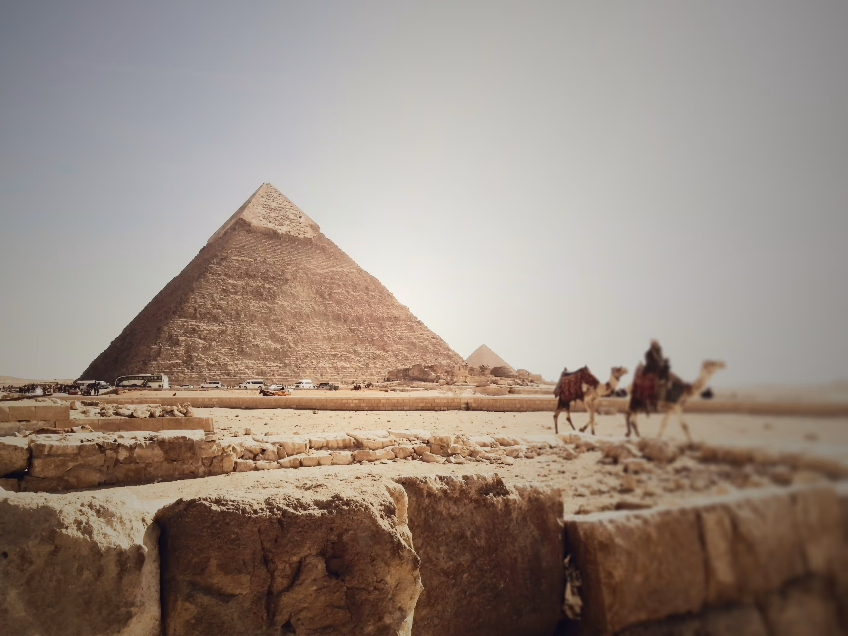 The Great Pyramid of Giza with camels and stone ruins in the foreground under a clear sky.