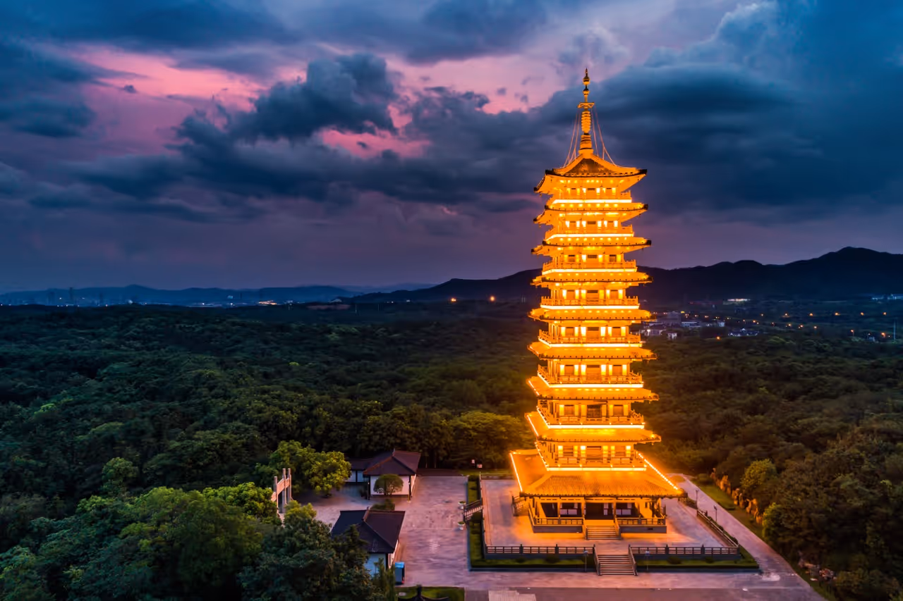 Illuminated multi-story pagoda glowing at dusk surrounded by forest and hills under a cloudy sky.
