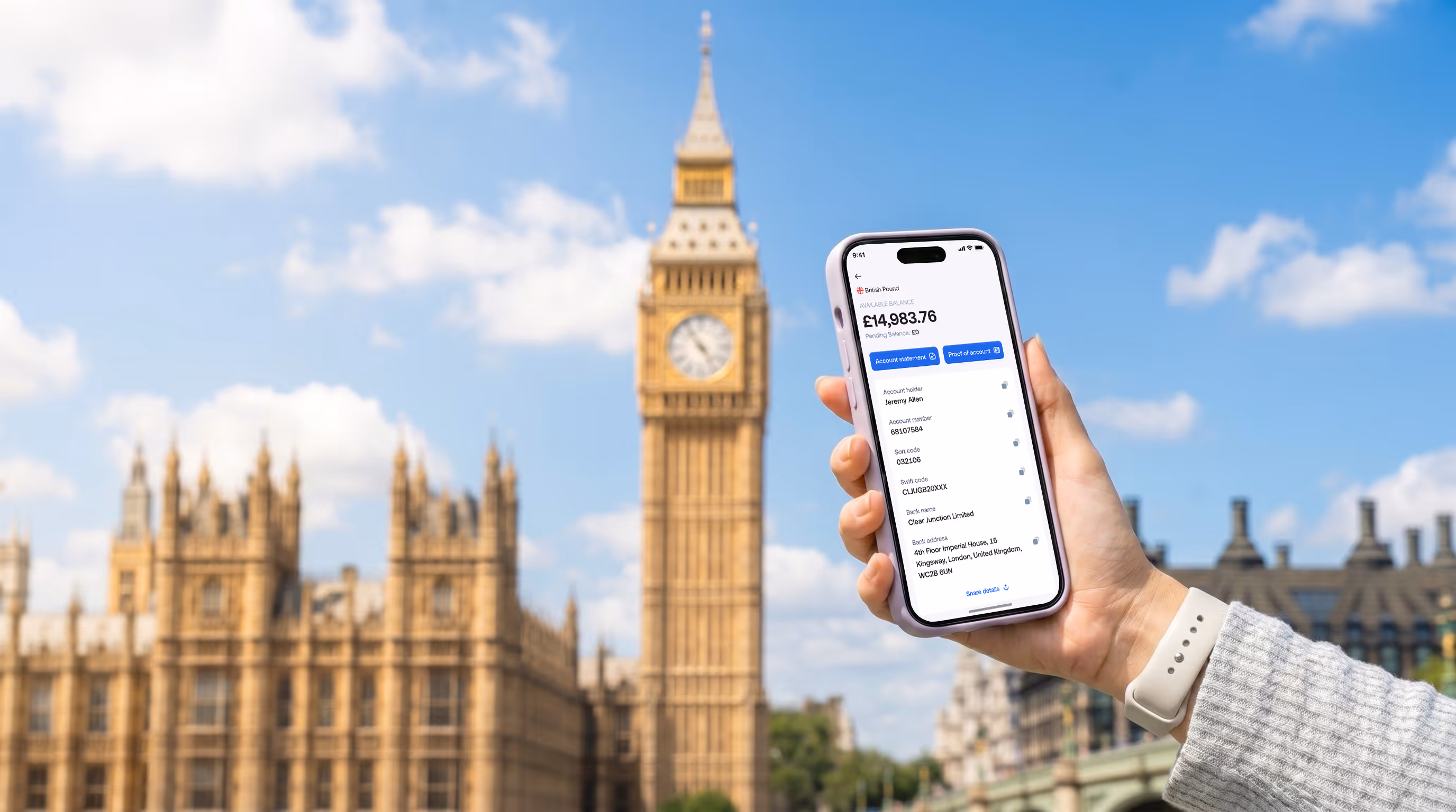 Hand holding smartphone showing a British Pound bank account balance of £14,983.76; Big Ben in background.