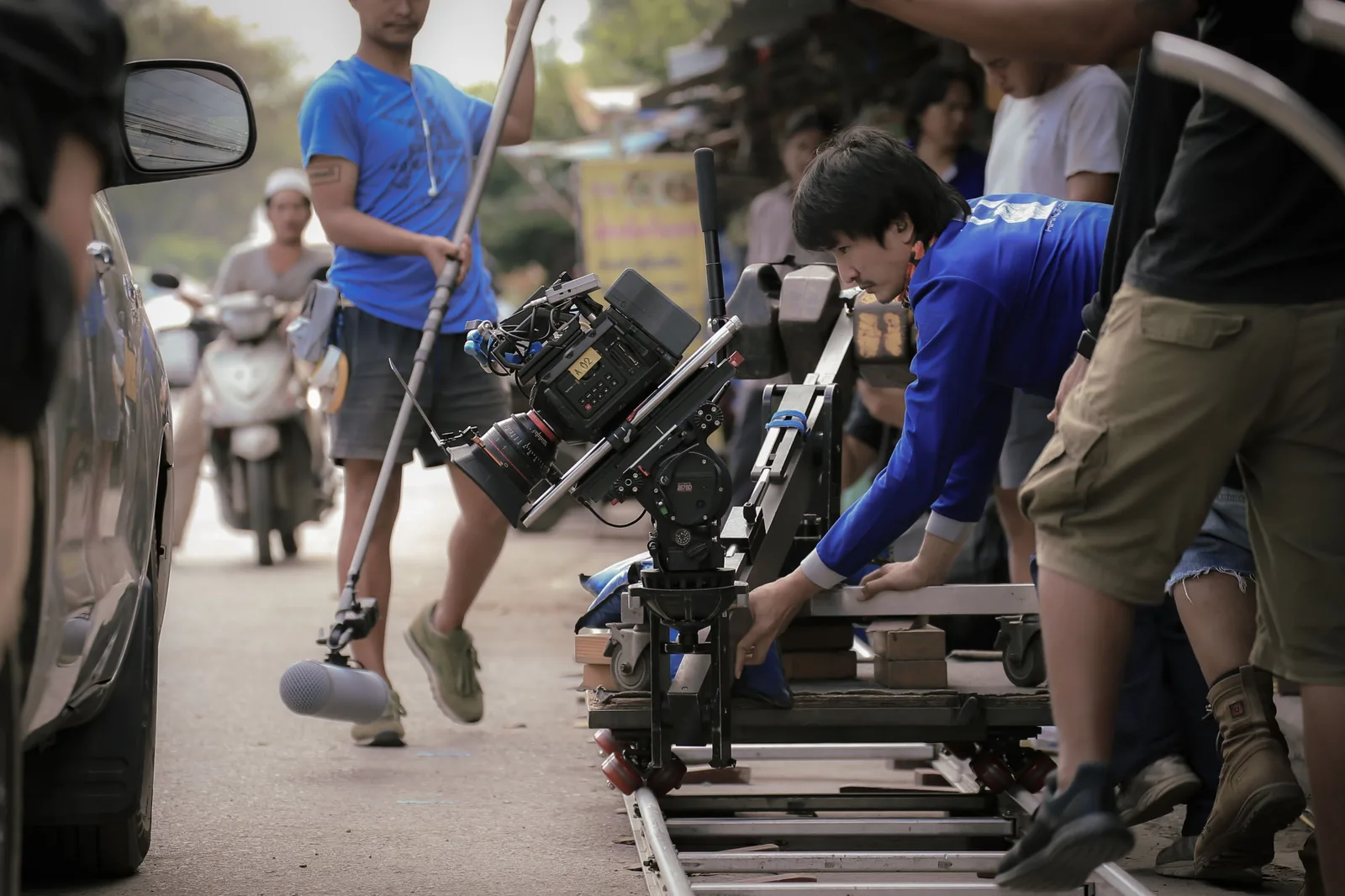 Grip positioning a camera dolly on track during an outdoor location shoot, with a cinema camera on a jib arm and a boom operator nearby