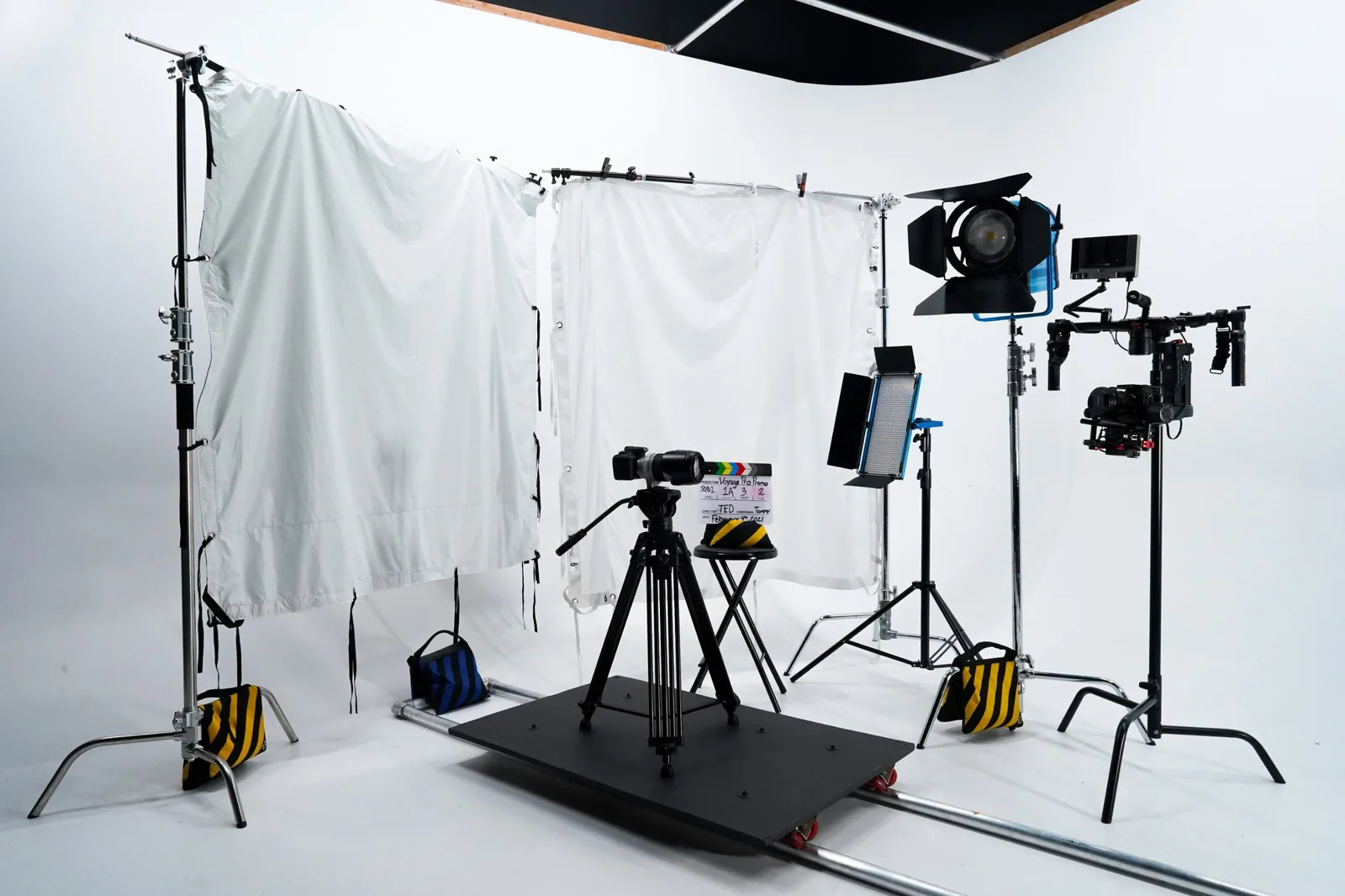 Studio setup with a camera on a tripod mounted to a dolly platform on track, beside a camera rigged on a handheld gimbal, with lights and diffusion frames