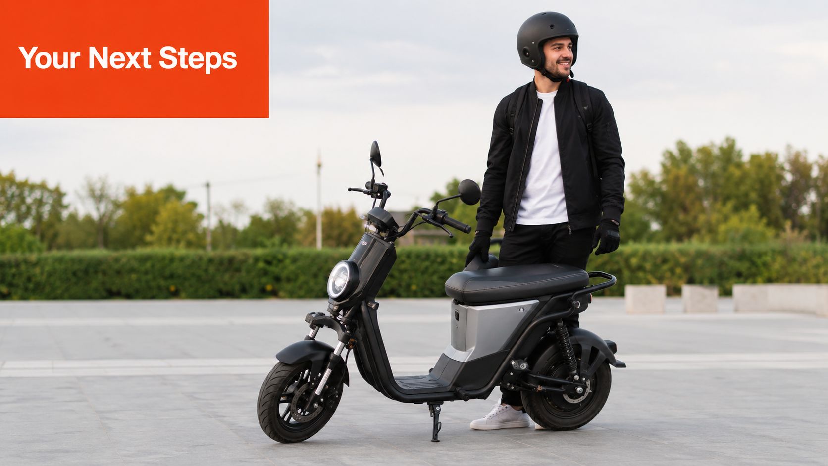 A young man wearing a helmet stands next to his sleek electric moped on a paved path.