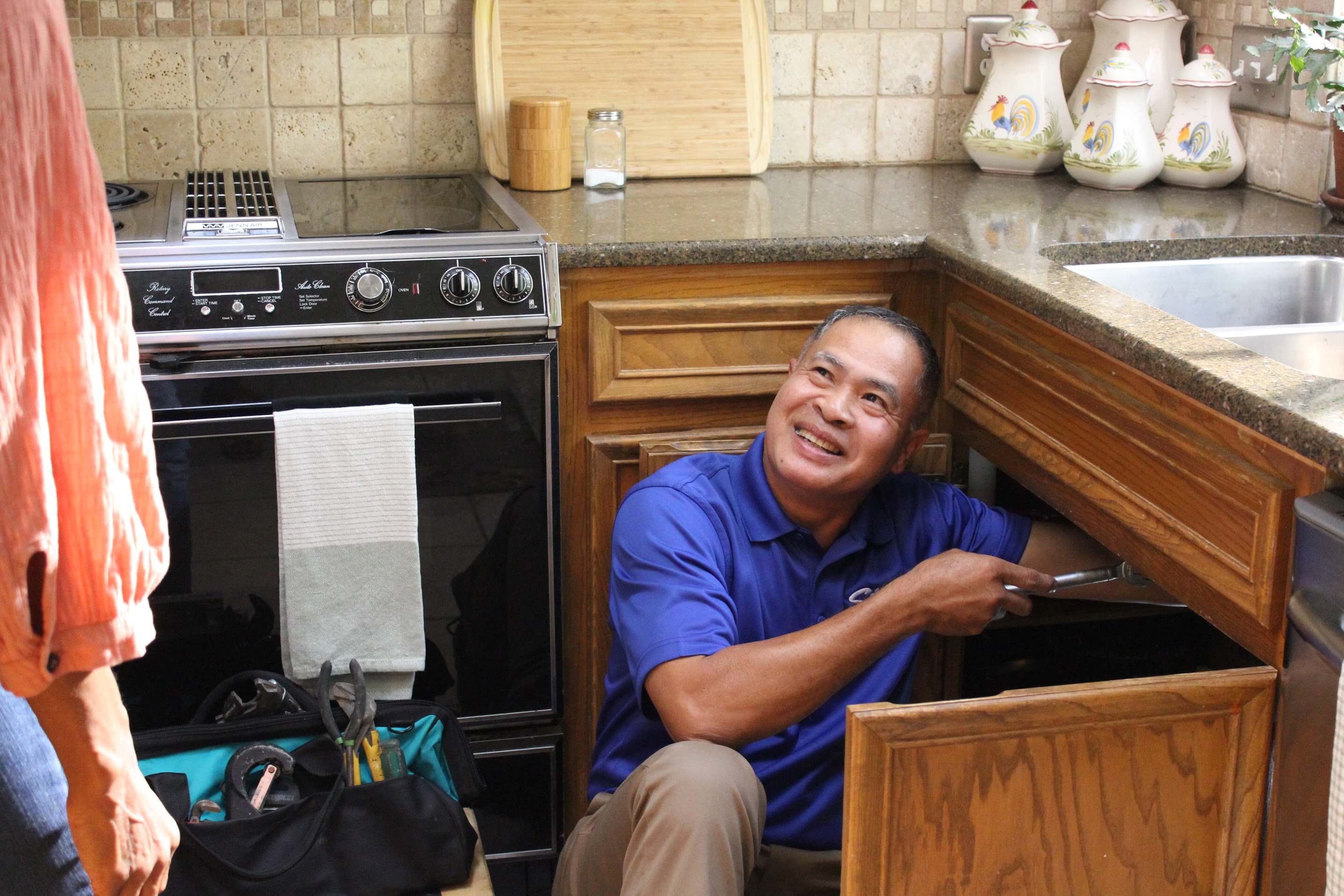 Plumber fixing kitchen sink pipe, smiling at customer, with tools visible on the floor.