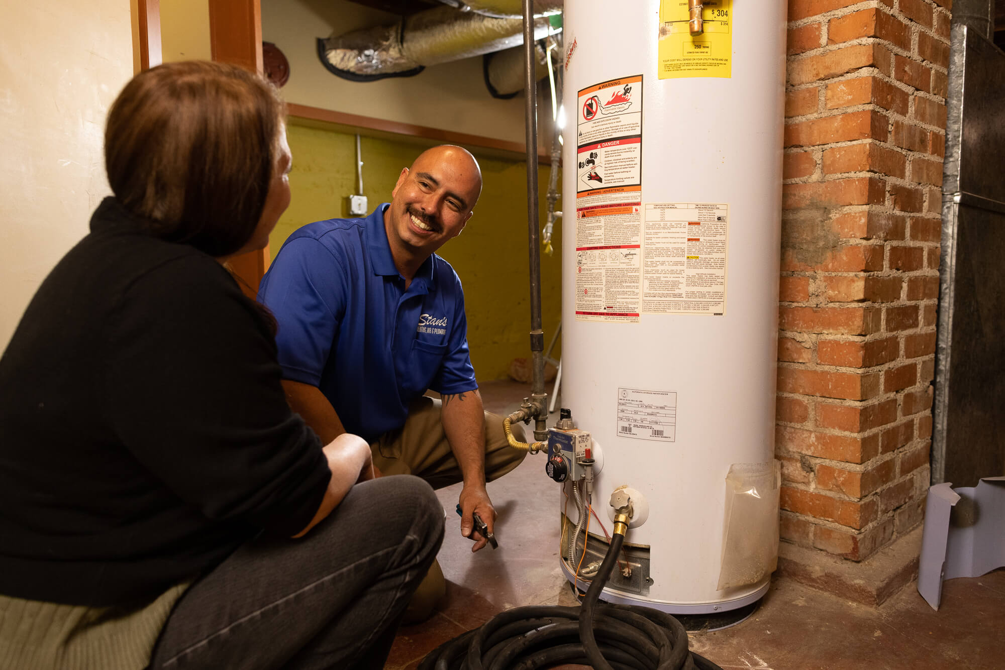 A technician in a blue shirt is inspecting a water heater while talking to a seated person in a basement.