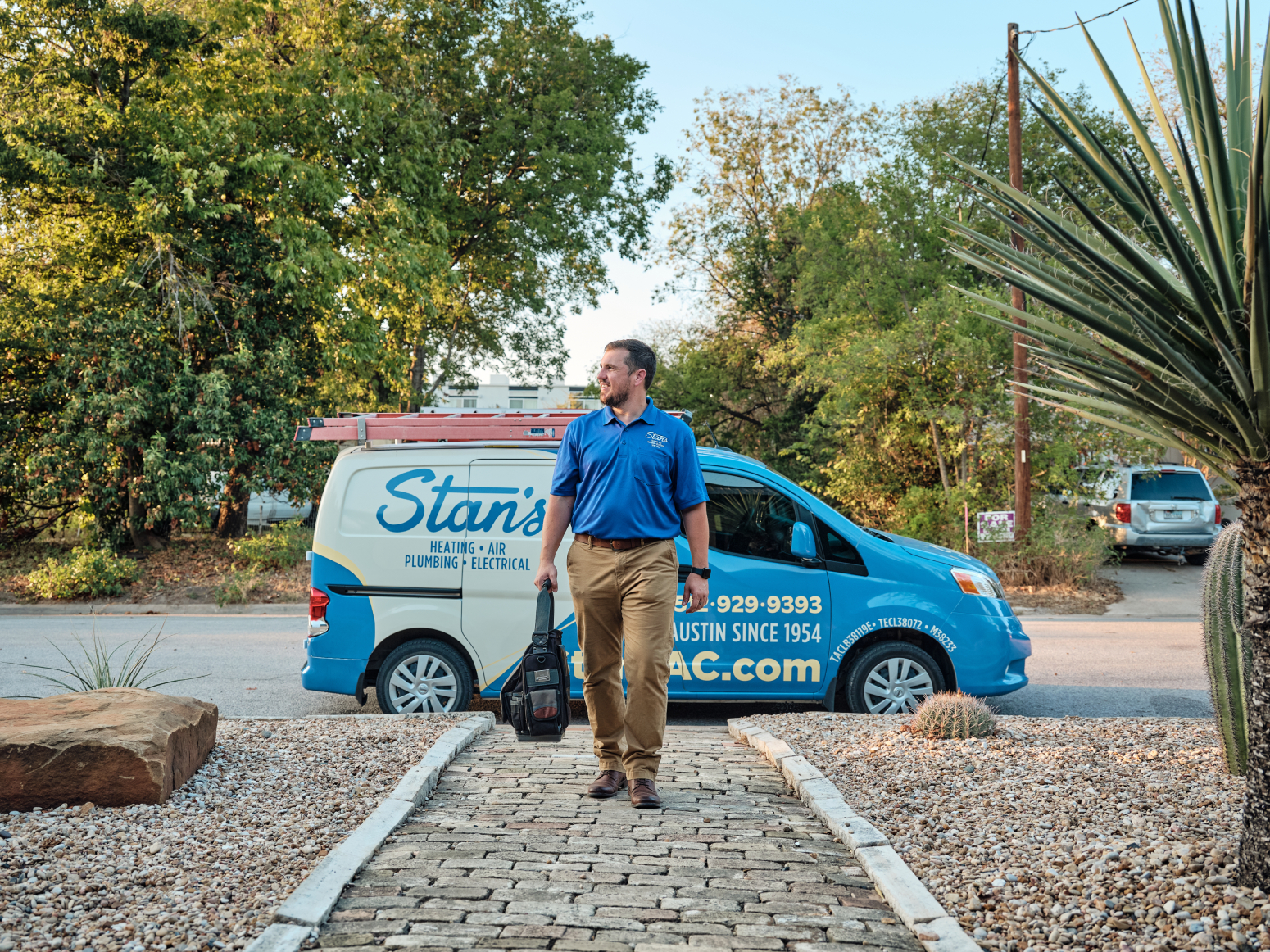 A person in a blue shirt and khaki pants stands outdoors in front of a blue van with trees in the background.