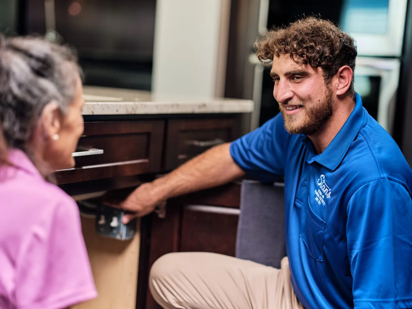 A male technician from Stan's is kneeling under a kitchen sink, working on plumbing while talking to a customer who is crouching beside him. They are engaged in a friendly conversation, and both are smiling.