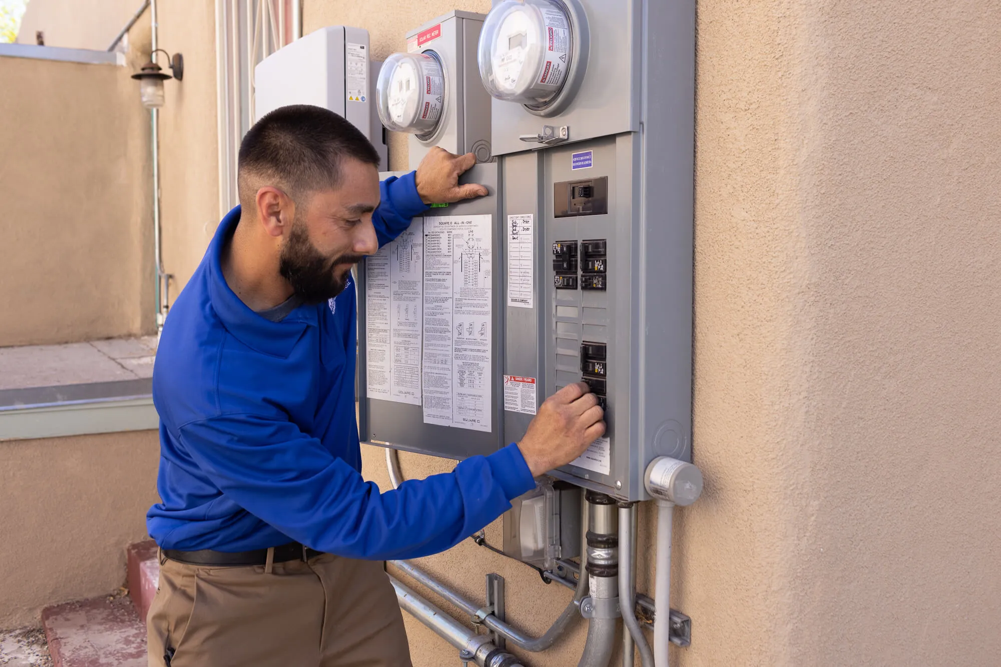 A Stan's Heating, Plumbing, and Electrical technician in a blue company polo shirt is adjusting the breakers on an electrical panel mounted on an exterior wall. The technician is focusing on the panel and has a slight smile.