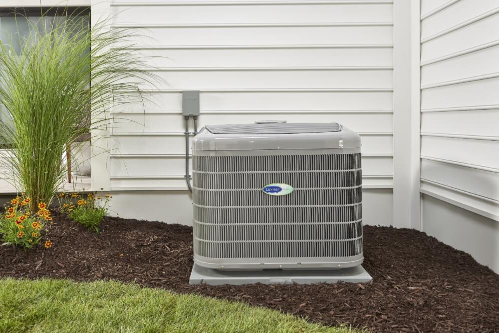 A high-efficiency air conditioning unit installed outside a house with white siding, surrounded by mulch and ornamental plants, representing a well-maintained HVAC system for home cooling.