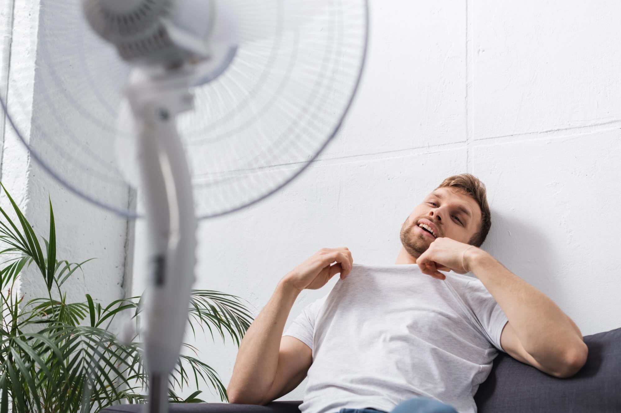Man relaxing on a sofa, cooling off with a fan in a warm room.