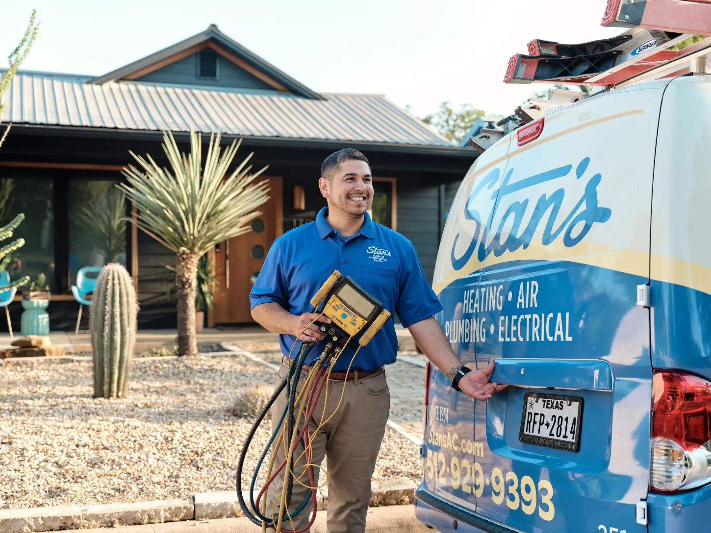 A male technician from Stan's is standing outside near a Stan's service van, smiling and holding a diagnostic tool with various cables. The background features a modern house with desert landscaping, including cacti and succulents.