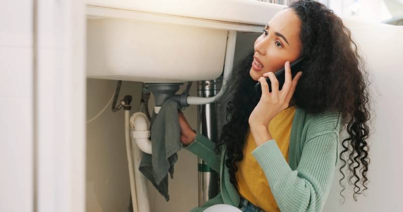Woman on phone inspecting sink plumbing.