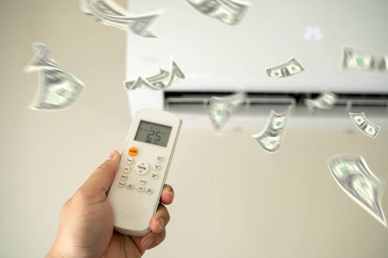 Hand holding AC remote with floating dollar bills.
