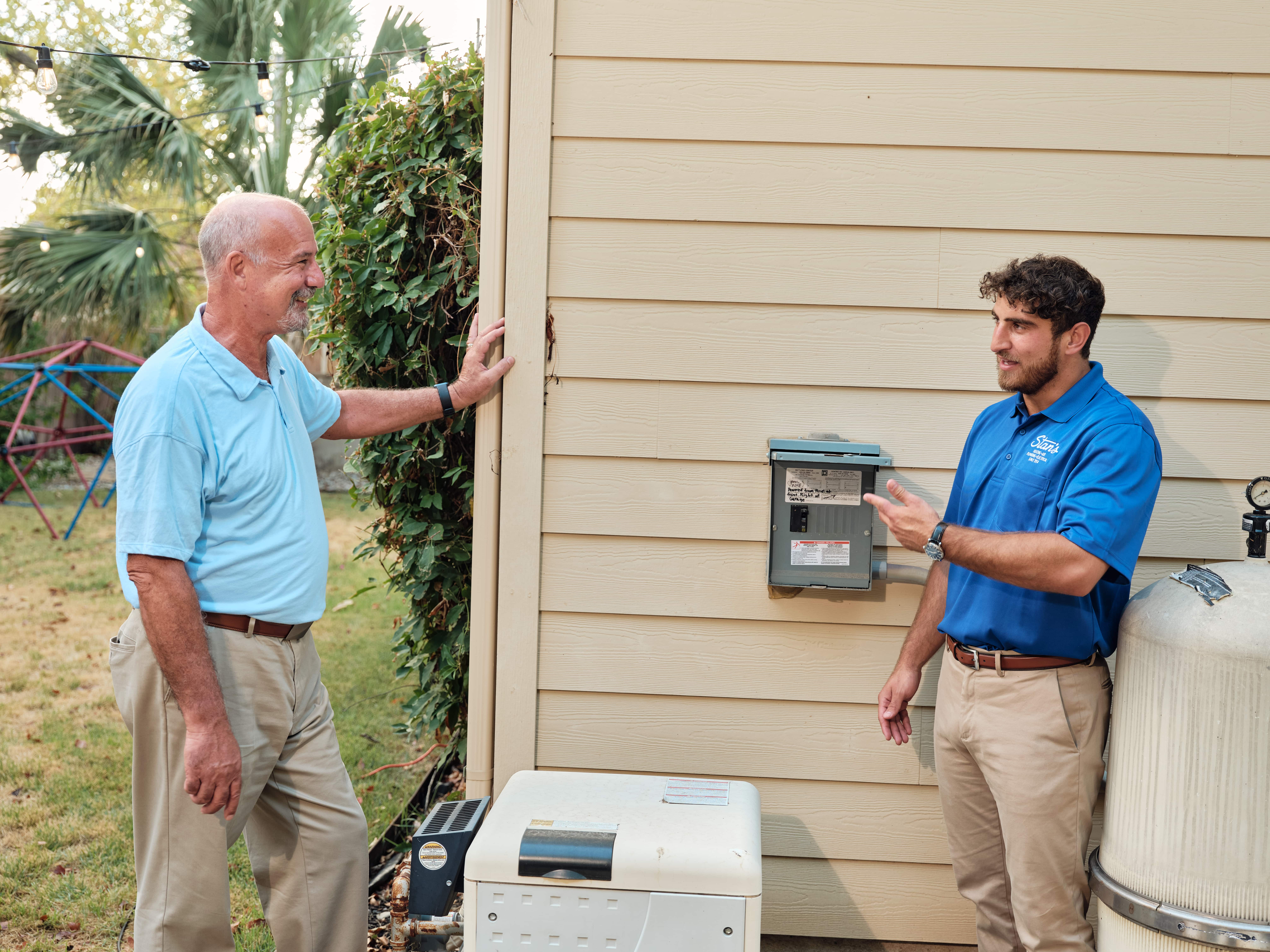 Two men in blue shirts discussing outdoor electrical panel.