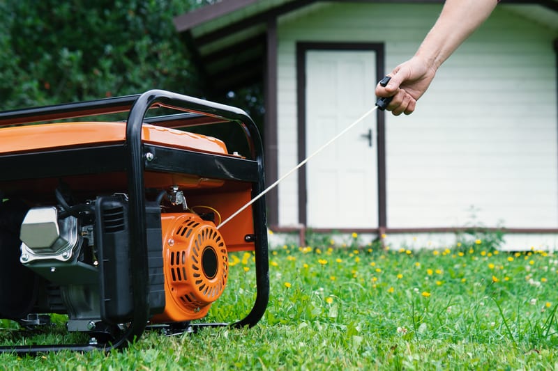 Person starting an orange portable generator near a small building.