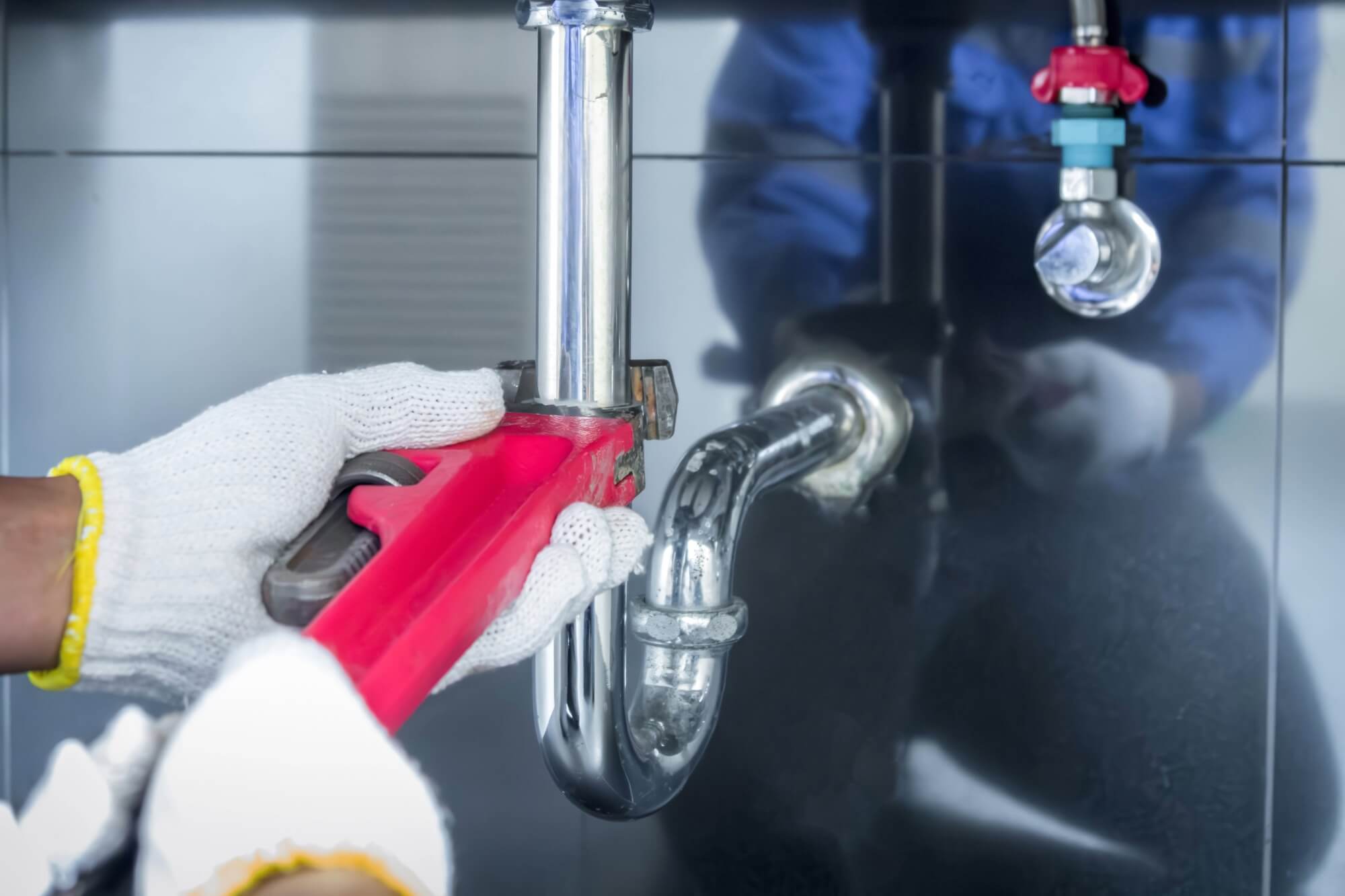 Plumber fixing a kitchen sink pipe with a wrench, wearing protective gloves and uniform.