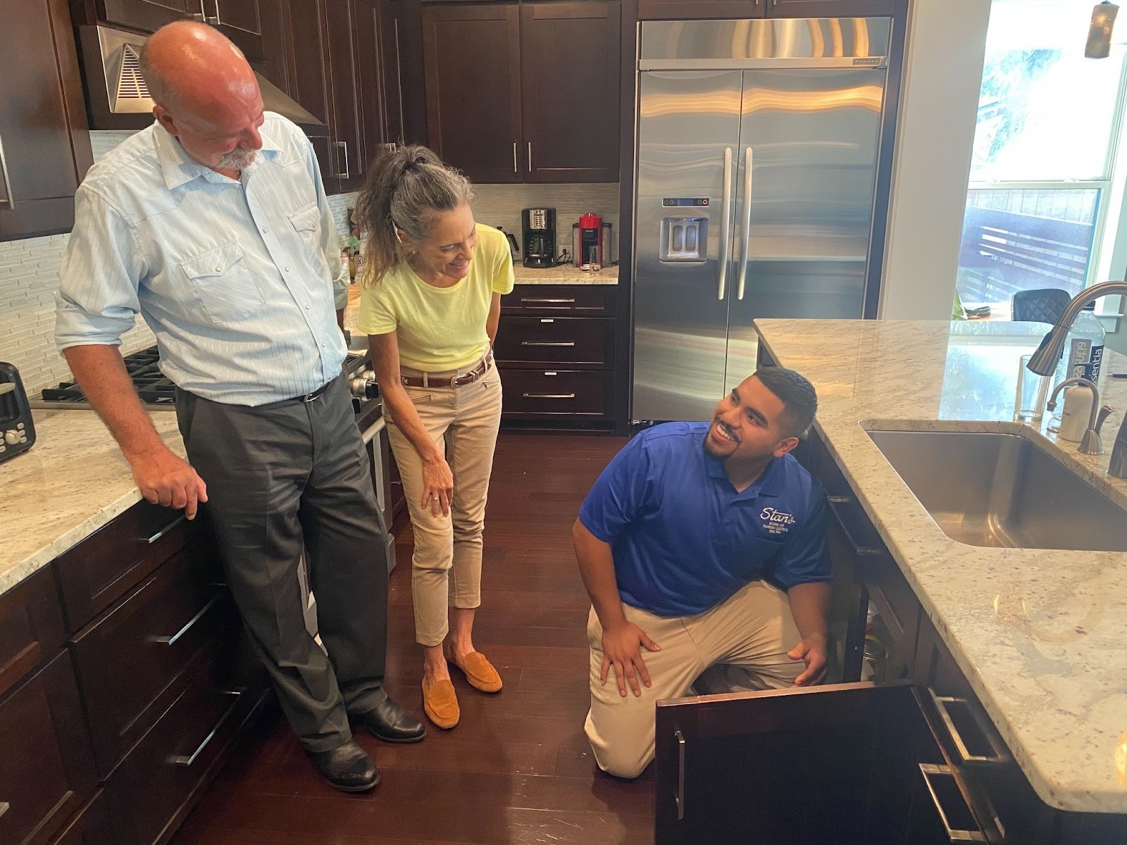 Technician in a blue Stan’s shirt talking with homeowners while checking plumbing under a kitchen sink