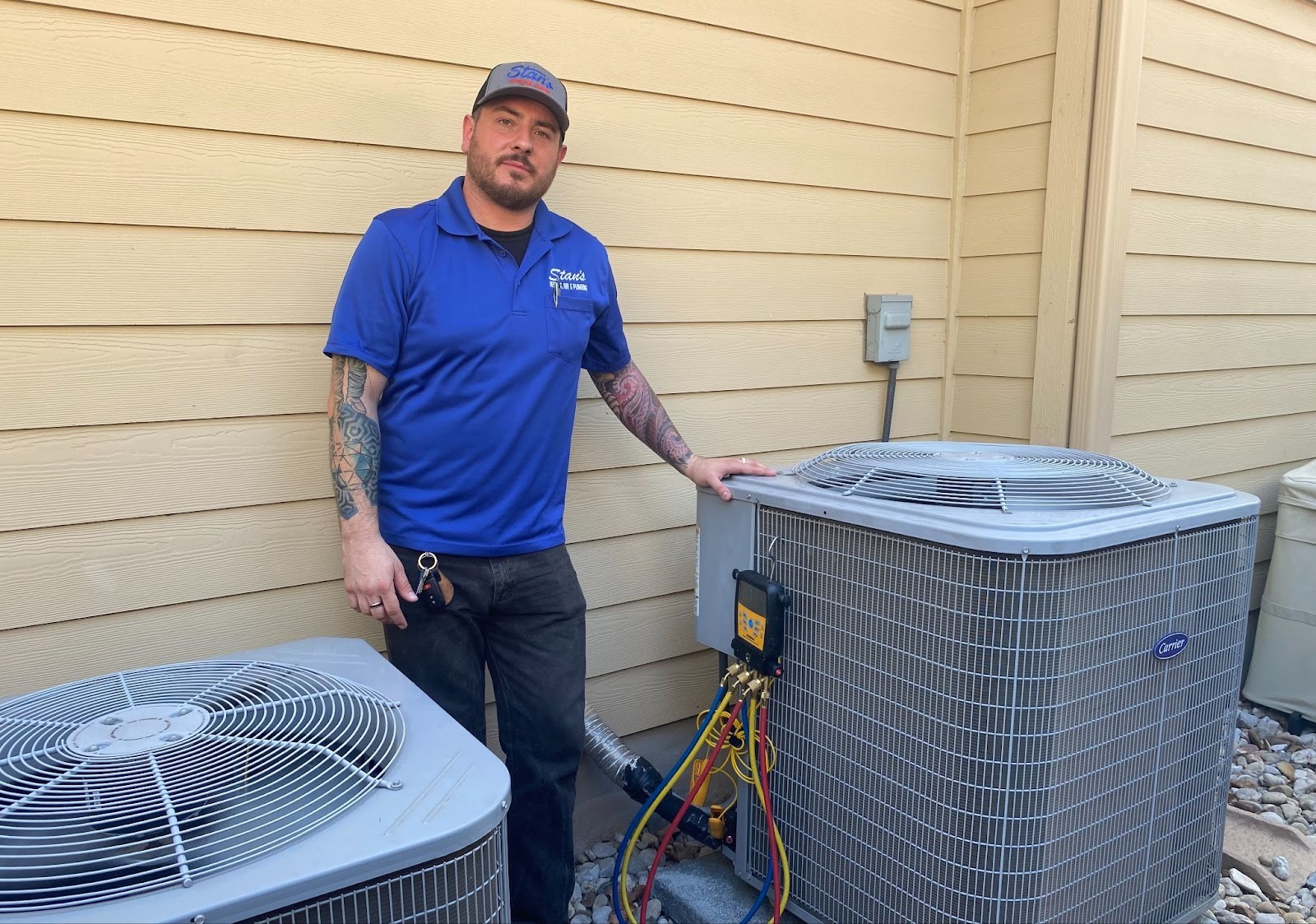 Technician in a blue Stan’s shirt standing next to outdoor air conditioning units during maintenance.