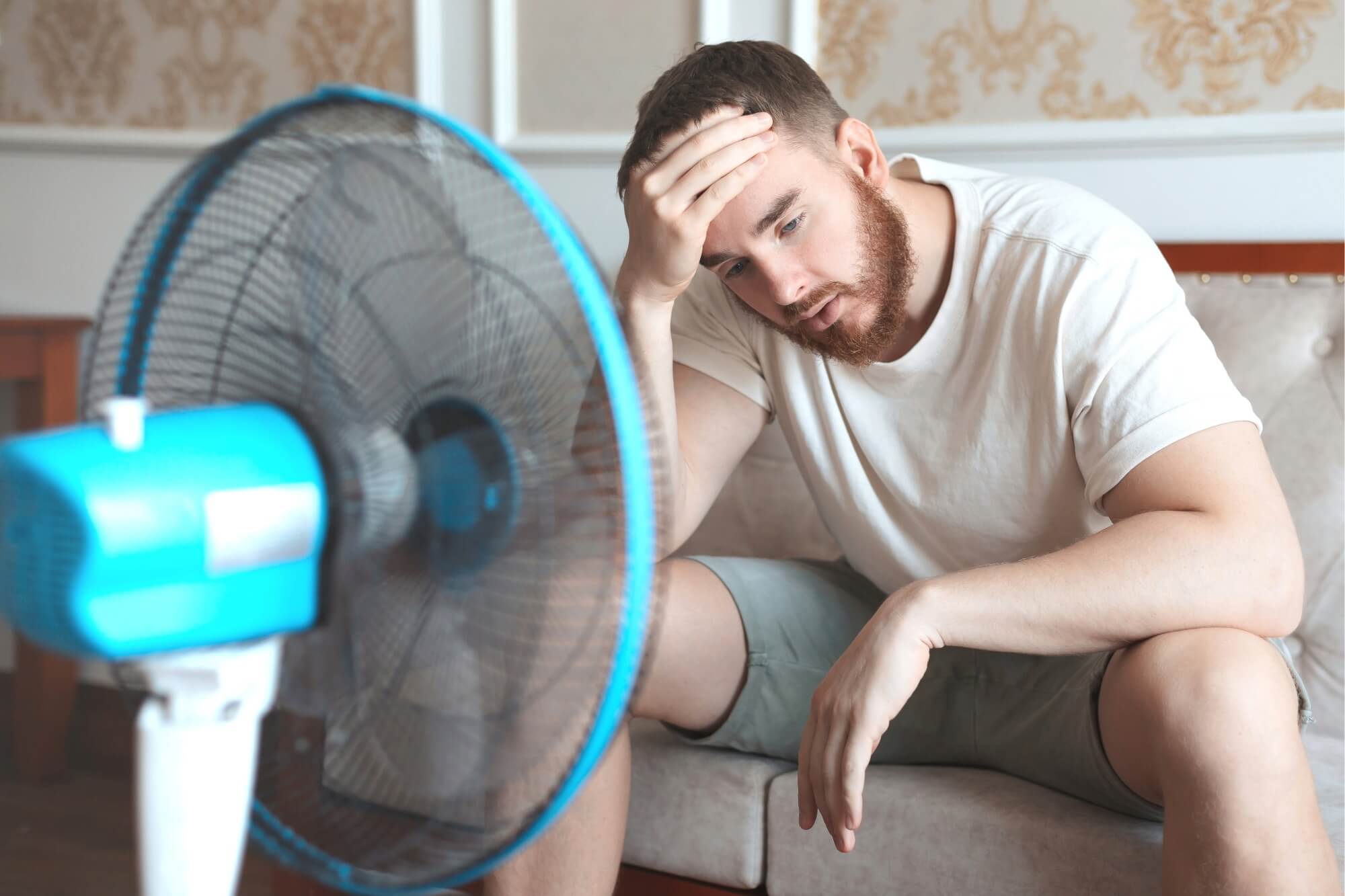 Man sitting on a couch, looking tired, in front of an electric fan during hot weather.