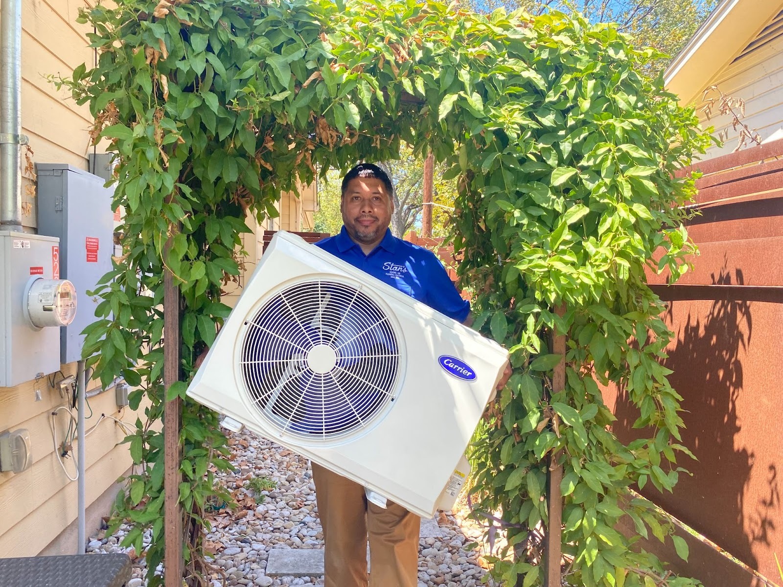 Technician in a blue Stan’s shirt holding an outdoor Carrier air conditioning unit near a house.