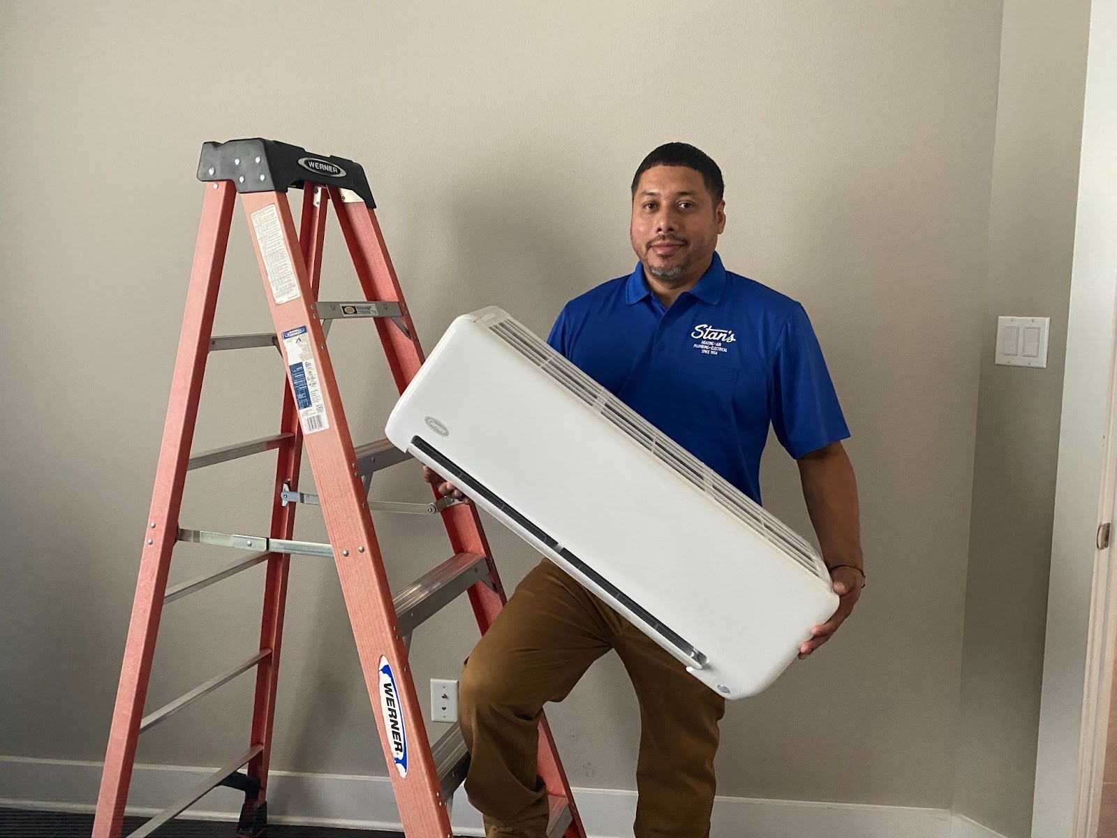 Technician in a blue Stan’s shirt holding an indoor AC unit next to a ladder, preparing for installation.
