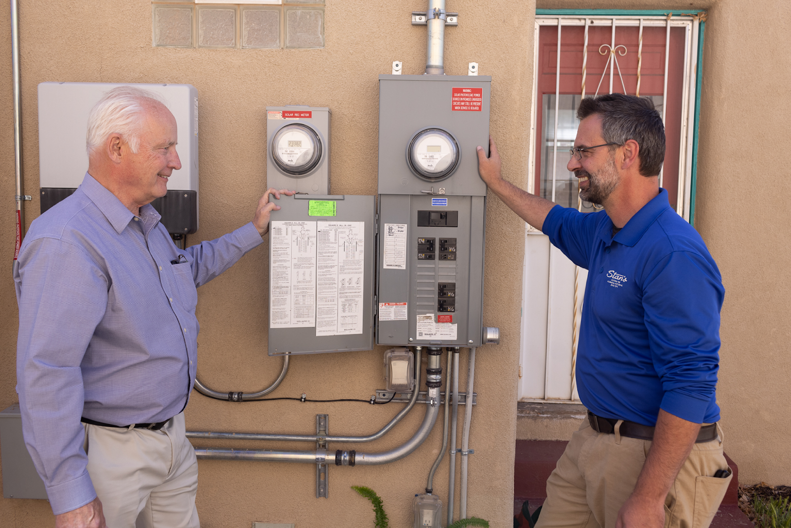 Technician in a blue Stan’s shirt discussing an outdoor electrical panel with a homeowner.