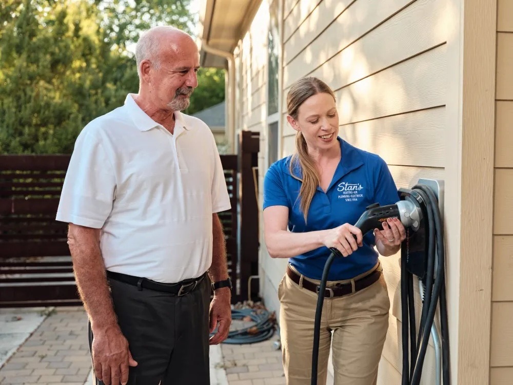 Alt img txt: Technician showing a homeowner how to use an electric vehicle charging station installed on a house exterior.