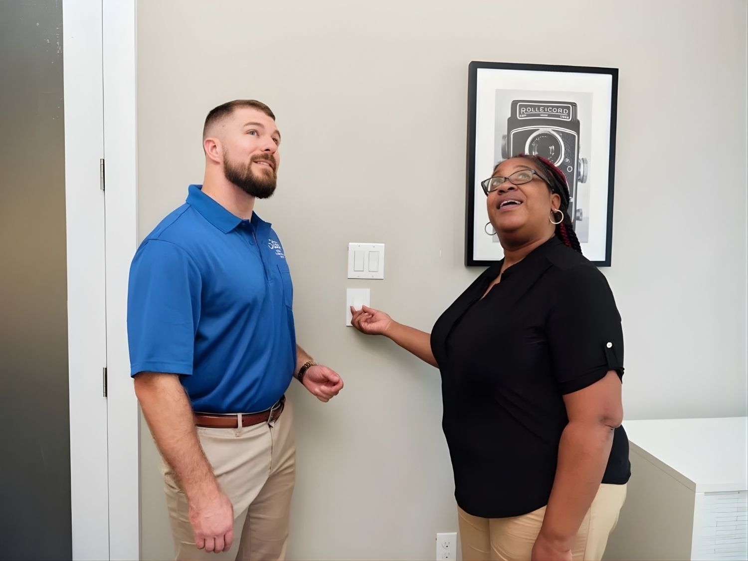 A man in a blue polo and a woman in a black shirt stand by a light switch on a wall, smiling and looking up, possibly discussing electrical features; a framed black and white photo of a vintage Rolleicord camera hangs nearby.