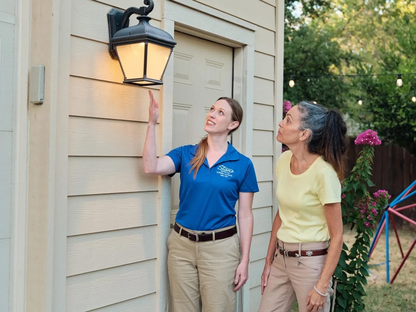 Technician in a blue Stan’s shirt showing a homeowner an outdoor wall light fixture on the side of a house.