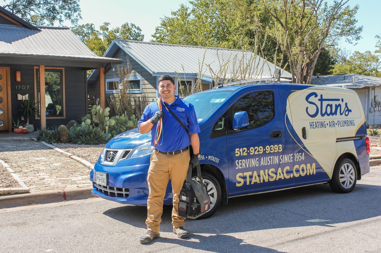 Technician standing in front of a Stan’s Heating, Air & Plumbing service van, holding tools and smiling outside a residential home