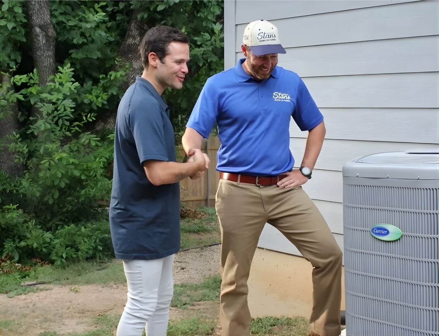 Two men standing outside near a house, one wearing a blue "Stan's" shirt and cap, likely an HVAC technician, discussing or inspecting a Carrier air conditioning unit.