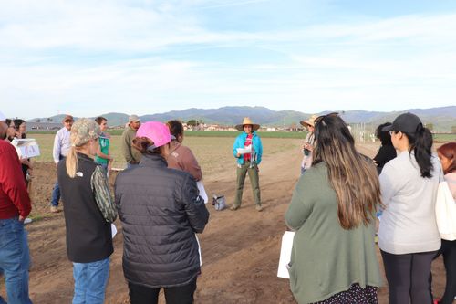 A group of farmers stand around and learn while a lecturer speaks