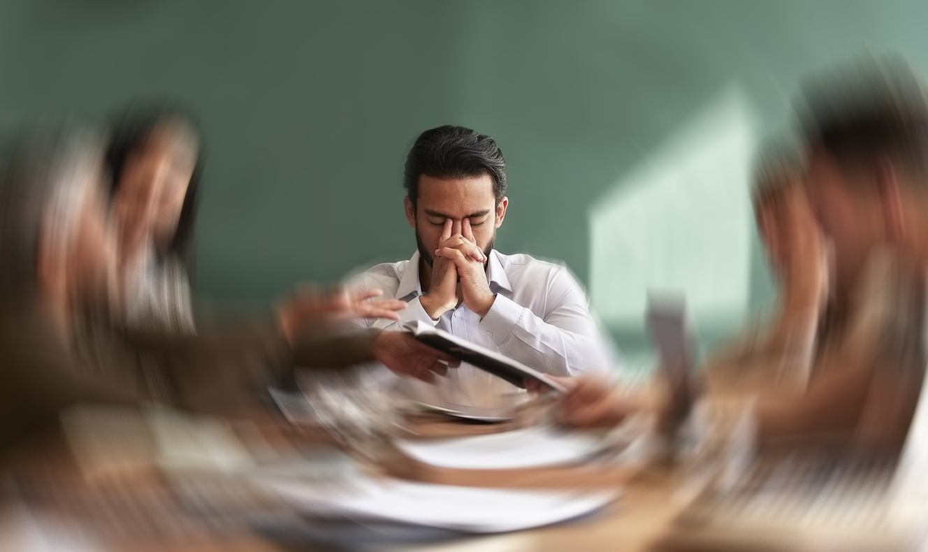 Man zit met gesloten ogen aan tafel terwijl wazige collega’s en documenten om hem heen bewegen, als beeld van werkdruk en mentale overbelasting.