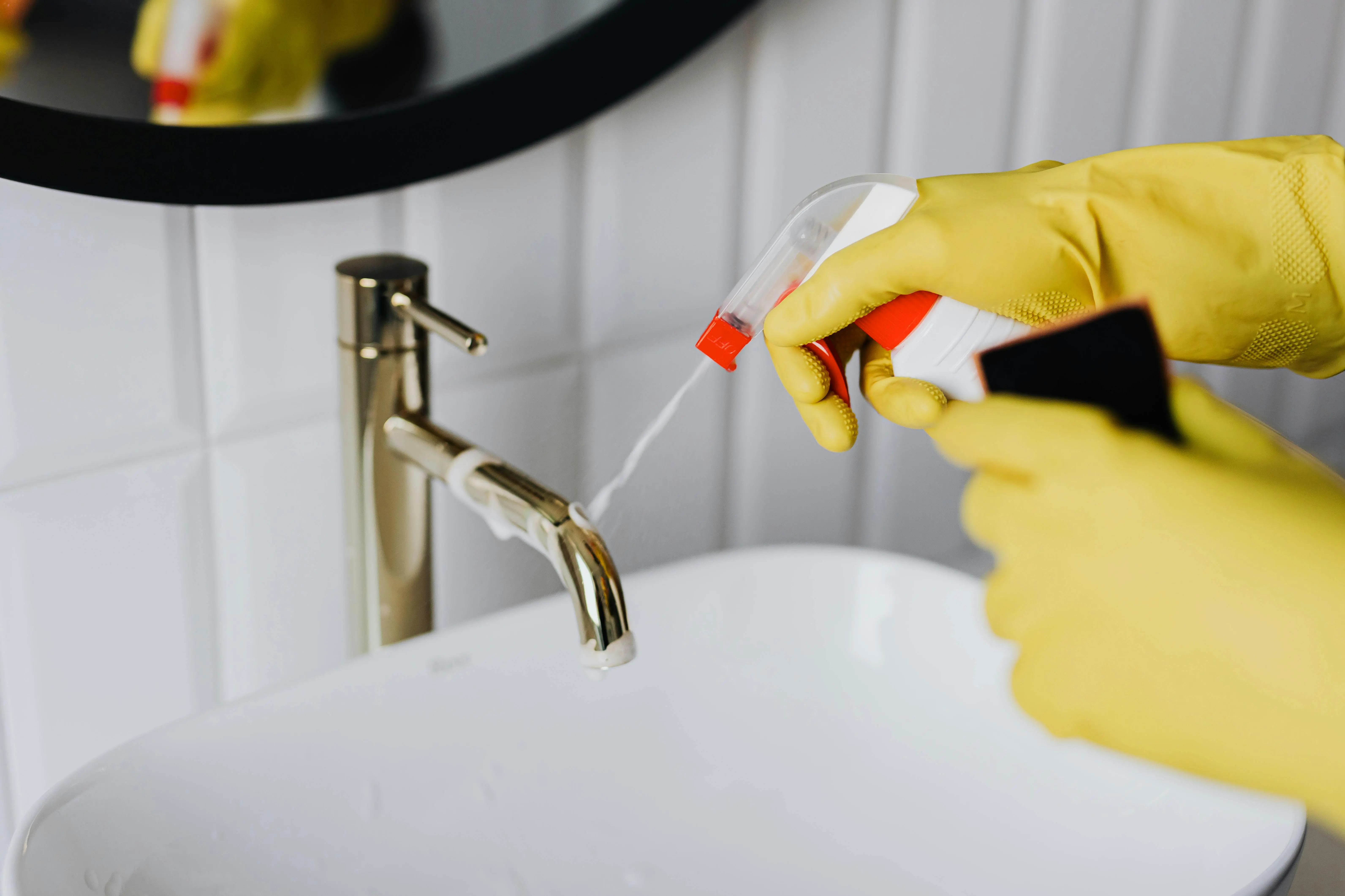 Close-up of gloved hands spraying and scrubbing a bathroom sink faucet, illustrating the benefits of recurring professional cleaning services in North York homes.