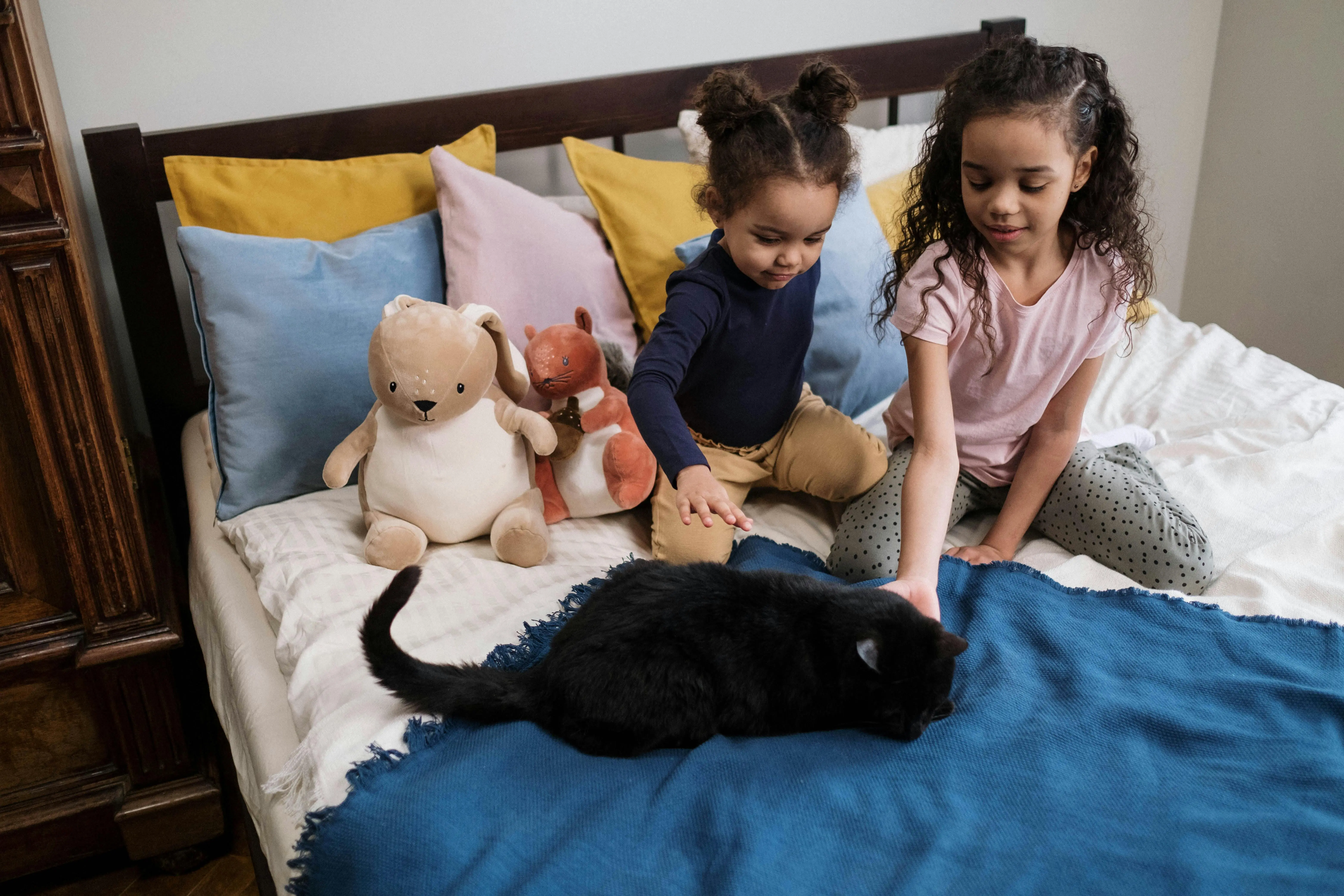 Two children sitting on a neatly made bed with stuffed animals and a black cat, highlighting the importance of weekly cleaning for families with kids and pets in Brampton.