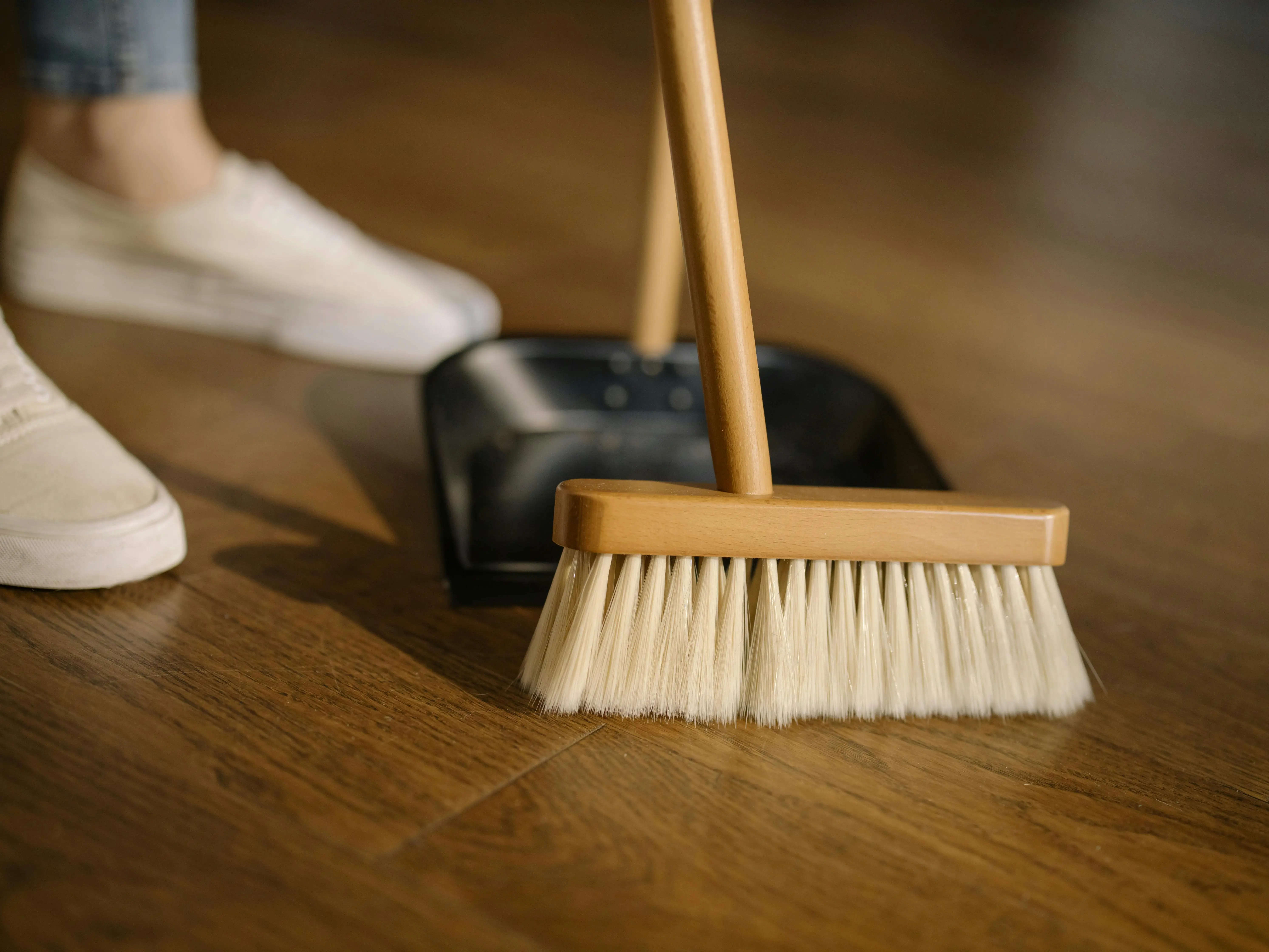 Person sweeping hardwood floor with a broom and dustpan, representing the importance of deep cleaning floors to remove fleas, eggs, and larvae during an infestation.