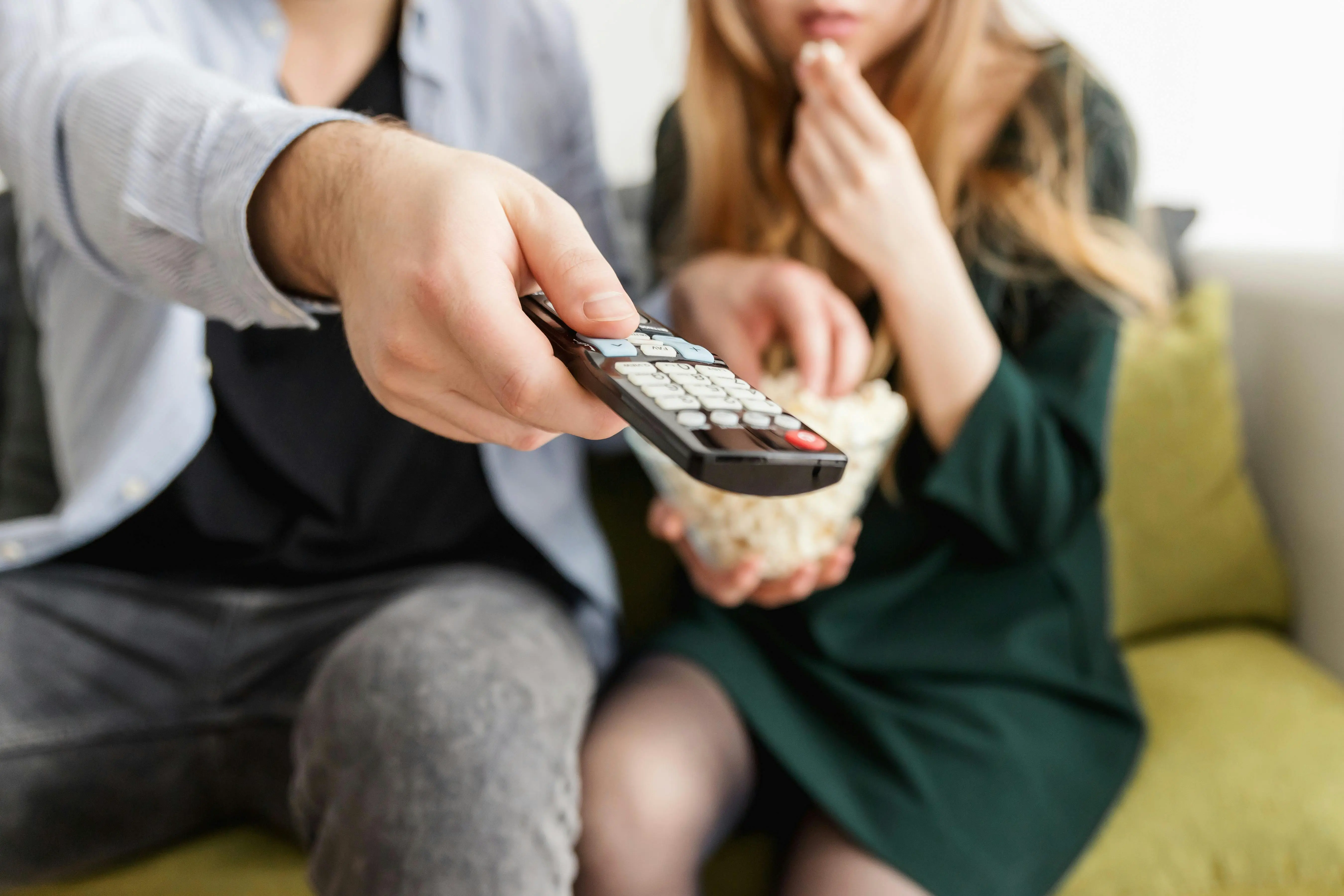 Close-up of a person holding a TV remote control while sitting on a couch with another person eating popcorn, highlighting how remotes are high-touch germ hotspots in the home.