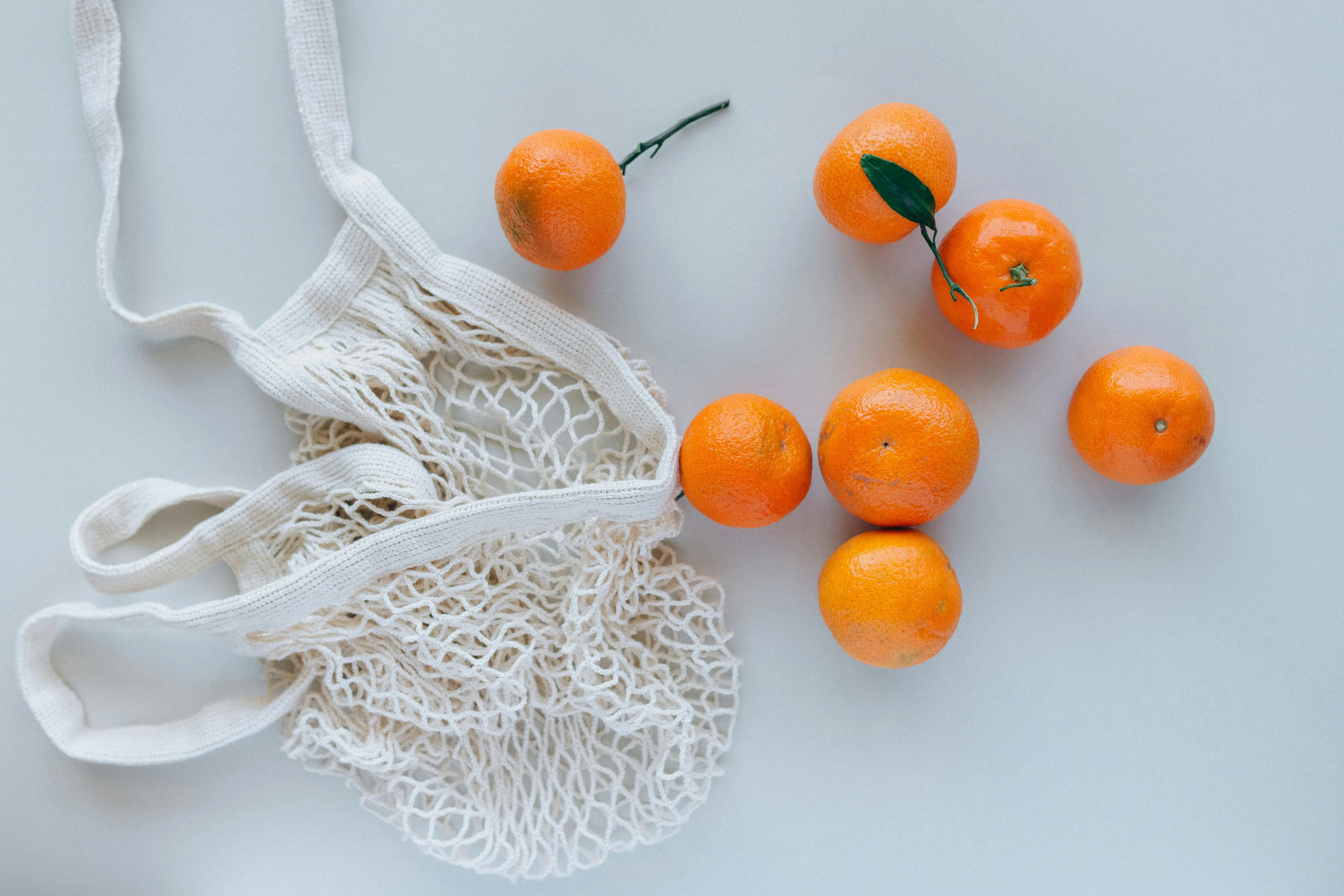 Mesh reusable grocery bag with fresh oranges placed on a white surface, highlighting the need for regular cleaning to prevent bacteria buildup.