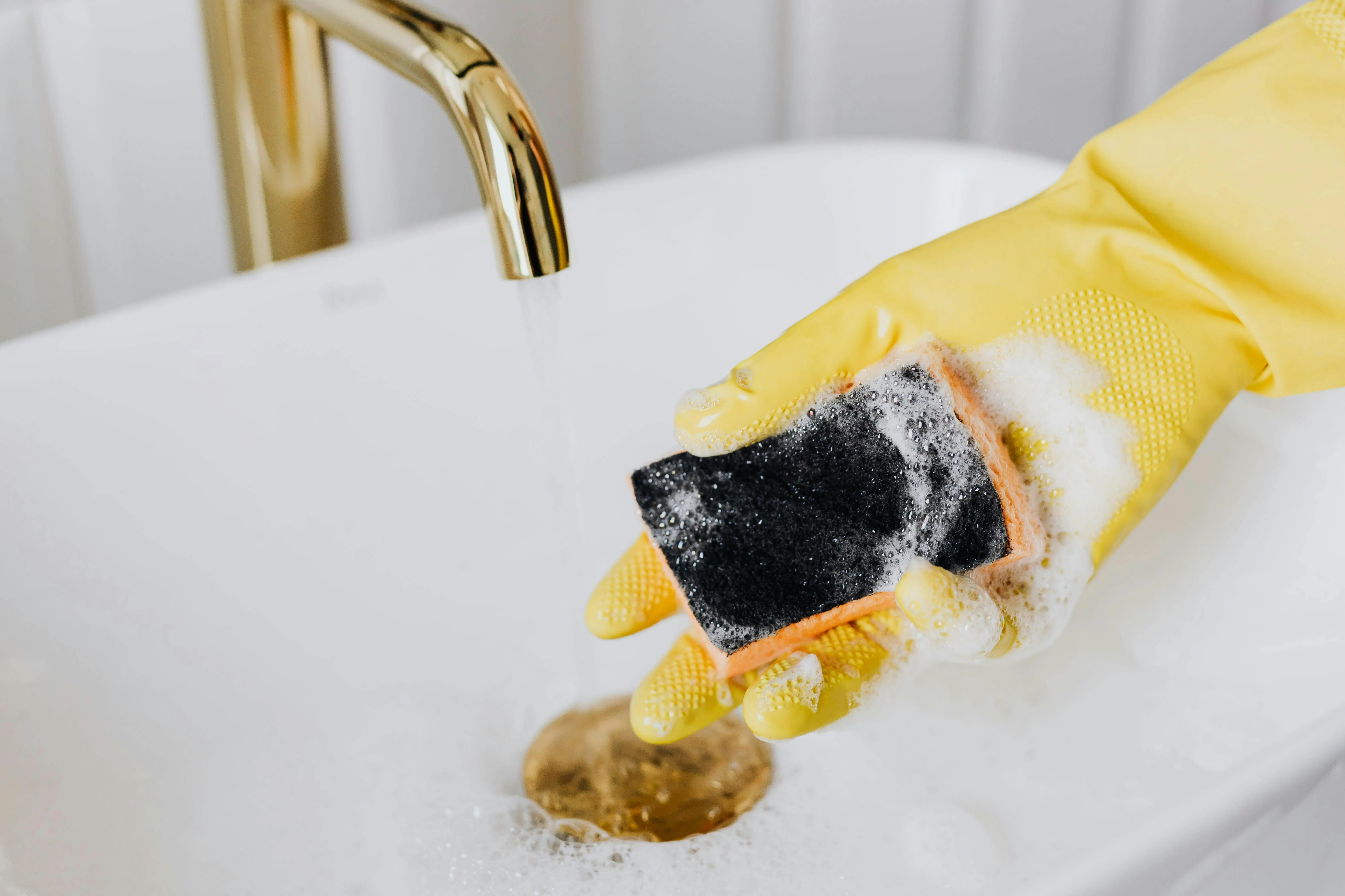 Hand wearing a yellow glove holding a wet kitchen sponge under running water in a sink.