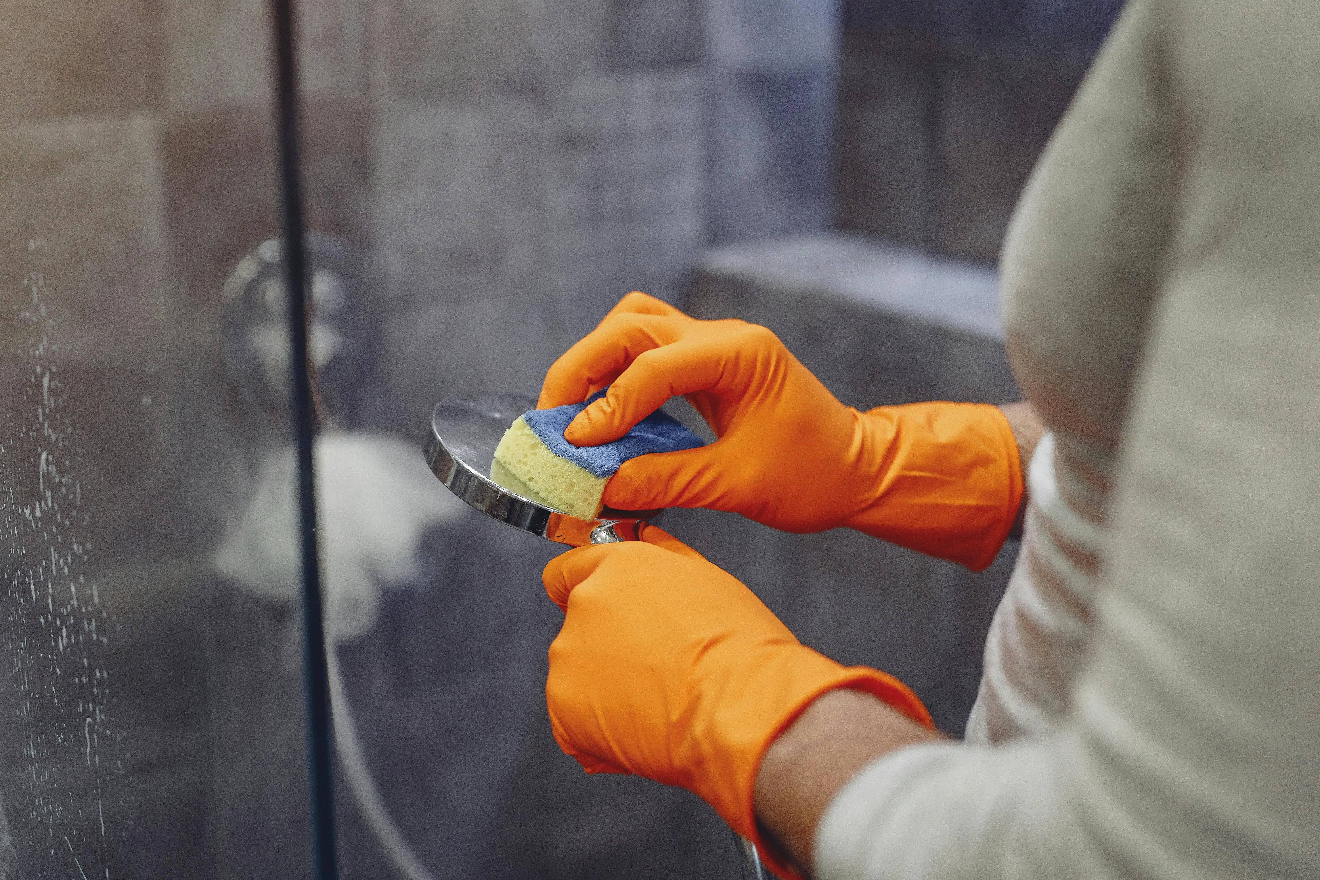 Close-up of hands in orange rubber gloves scrubbing mold on bathroom tiles with a sponge.