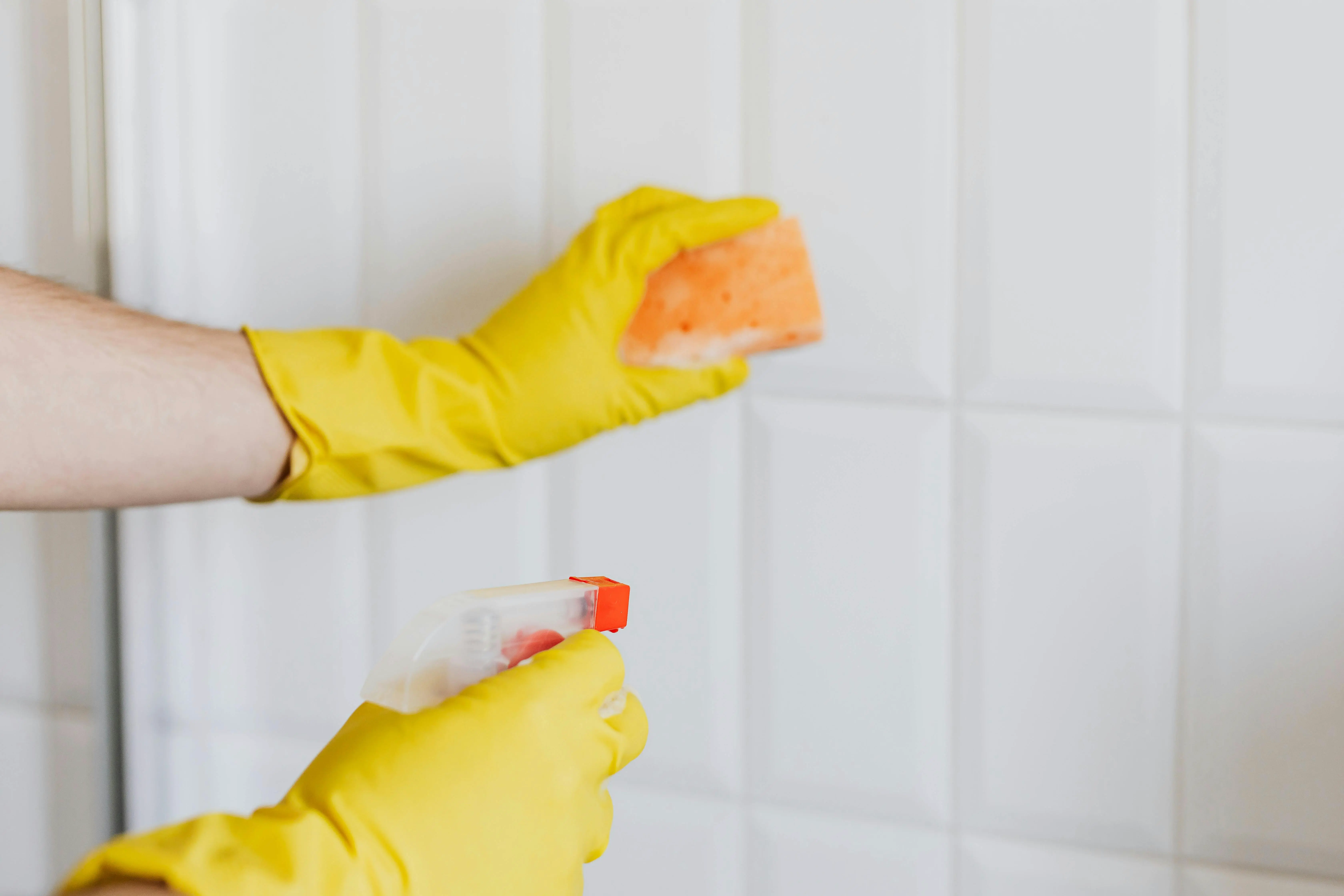  Person wearing yellow gloves cleaning tiled wall with spray bottle – Oakville family weekly cleaning routine for a healthier home.