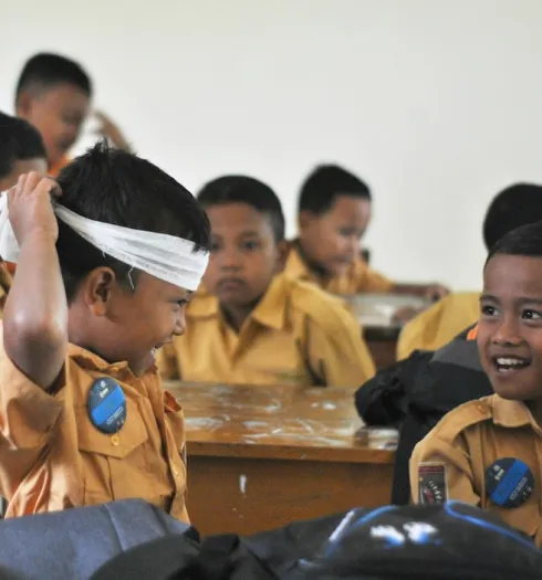 Young schoolboys in yellow uniforms sitting at desks, one boy playfully tying a white cloth around his head while smiling at another boy.