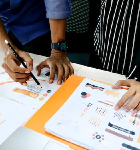 Two people reviewing and marking printed documents with charts and data, one holding a pencil.