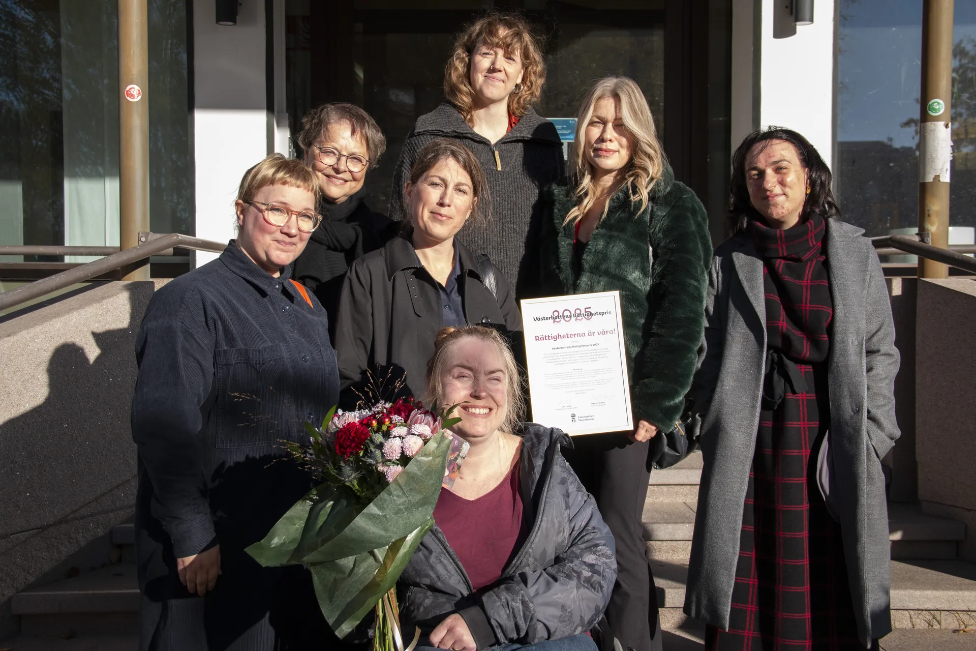 En grupp på sju kvinnor poserar utomhus på en trappa vid en byggnadsentré. En kvinna håller ett diplom med texten 'Rättigheterna är våra!' och en annan håller en blombukett. De firar mottagandet av Västerbottens Rättighetspris.