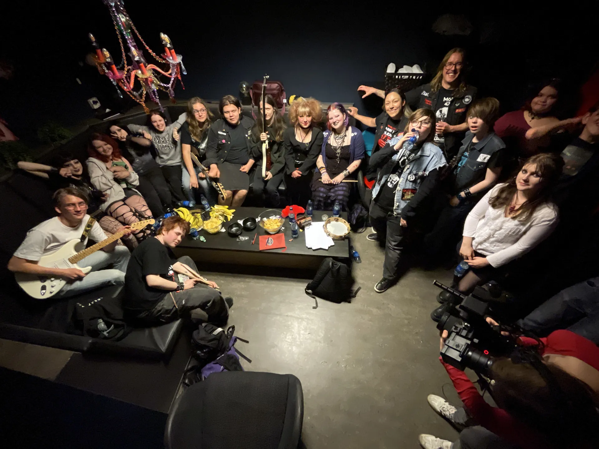 En grupp ungdomar och vuxna samlade i ett mörkt rum med musikinstrument som gitarrer och trumstockar, sittande runt ett soffbord med en färgglad ljuskrona i bakgrunden